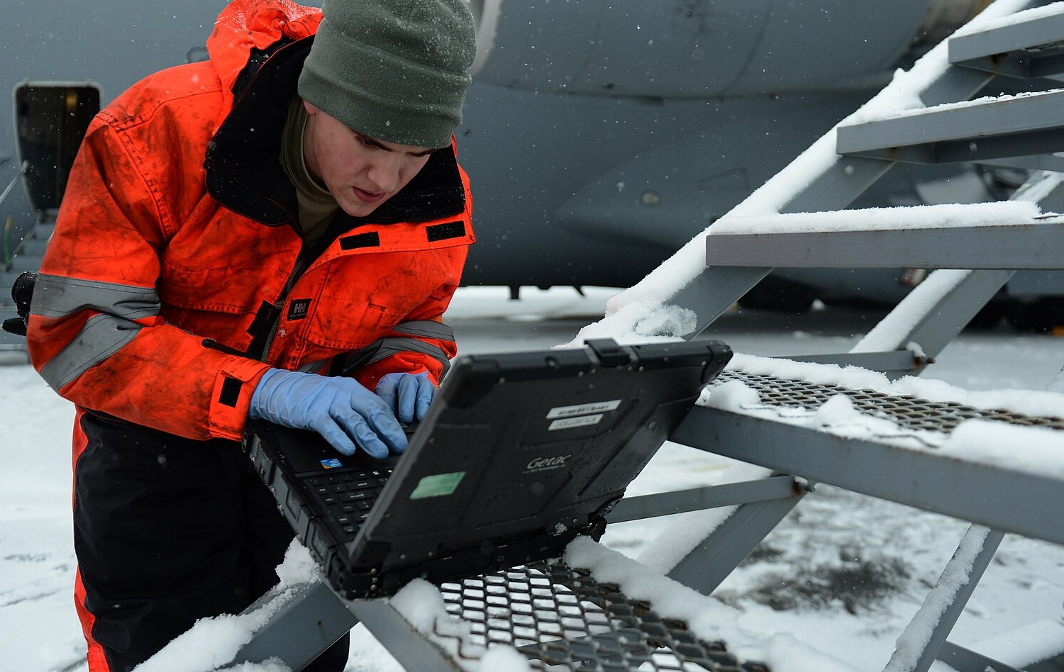 A technician in a work jacket kneels in the snow beside an aircraft and checks the procedure on a rugged laptop.